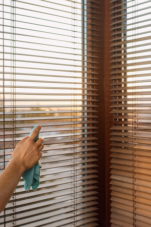 A person is cleaning wooden window blinds with a cloth while the sun sets outside, creating a warm...