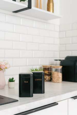 A modern kitchen countertop displays tea and coffee in metal boxes beside small plants and glass...