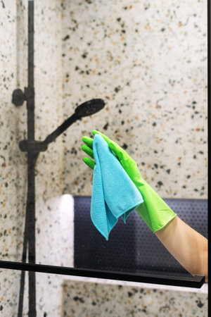 Cropped shot of a woman hand uses a blue cleaning cloth to wipe down the shower glass in a modern...