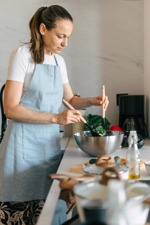In a modern kitchen, a woman is focused on mixing fresh vegetables in a salad bowl, promoting a...