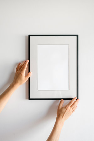 Woman hands is carefully adjusting an empty black picture frame against a white wall in a...