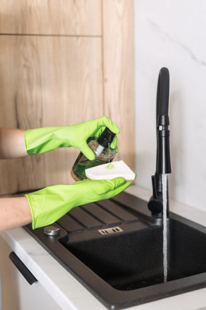 A person wearing green gloves uses a sponge and dish soap to clean a modern kitchen sink. The sleek...