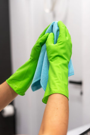 In a residential bathroom, a woman wearing bright green gloves is diligently cleaning with a blue...