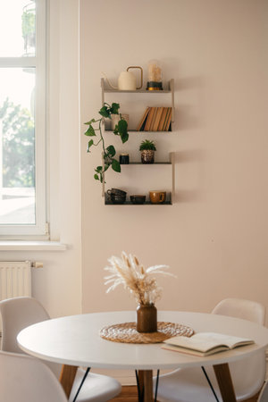 Reading at morning. Modern kitchen features a white table surrounded by minimalist chairs and a wall shelf adorned with plants and decorative items. Concepts of slow living at homeの写真素材