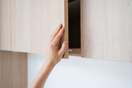 A hand reaches out to open a sleek wooden cabinet in a stylish, contemporary kitchen. This inviting space blends functionality furniture with modern aesthetics for a cozy home atmosphere.の写真素材