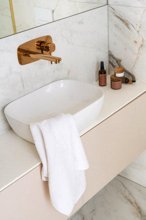 A well-designed bathroom features a stylish white sink and bronze faucet. Soft towels drape over the edge, alongside various self-care products, creating a serene morning routine atmosphere.の写真素材