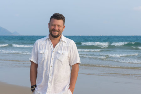 Portrait of a man with a middle beard in a white shirt, on the beach, on the background of the seaの写真素材