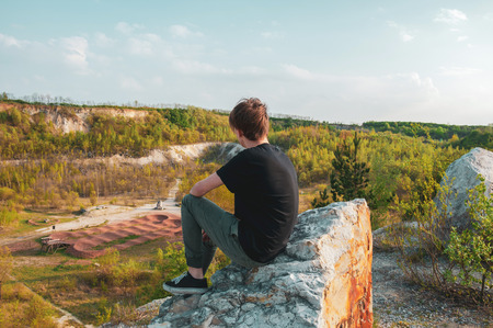 Young man tourist in black tshirt sitting on the giant rock, on the top of the mountain, during spring with warm colorsの写真素材