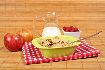 A bowl of cornflakes, milk, two apples and raspberries on bamboo mat