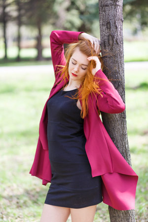 Red-haired girl posing near a tree in red clothes