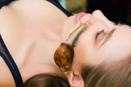 A large snail Akhatina crawls on the face of a young woman and rejuvenates her skin. The concept of unusual care and rejuvenation of the skin of the face.の写真素材