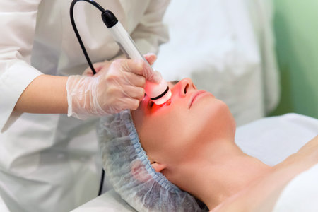 A young woman in a spa salon is having a face lift. The beautician holds the hand piece of the...
