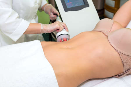 Young beautiful woman lies on a radiofrequency lifting procedure. The beautician of the spa salon holds the maniple over the abdomen.の写真素材