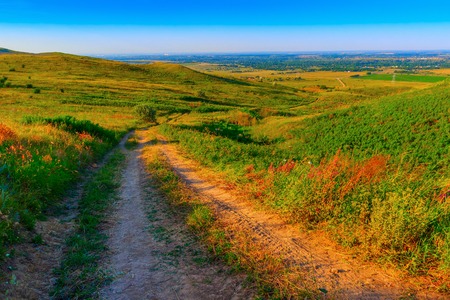 The twisting dirt road downhill the hill. On the parties, on slopes of hills, green herbs and bushes.の写真素材