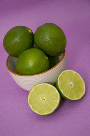 A white bowl of fresh green limes on a bright purple background with the main point of focus on the two cut limes in front of the bowl.の写真素材