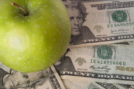 A green apple sits on top of a pile of $20 bills to illustrate the cost of education, food, or...
