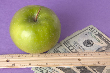 Conceptual image of a bright green apple with a ruler, and American currency on a vibtrant purple background to represent the cost of education.の写真素材