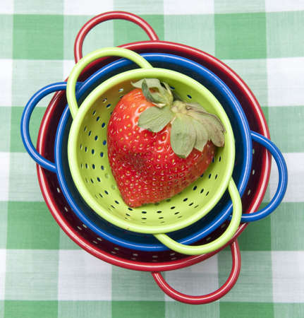 Trio of colorful colanders topped with a ripe strawberry on a green picnic blanket background.  Healthy summertime eating.の写真素材