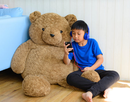 Little asian boy in headset using smartphone with teddy at home