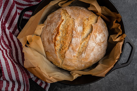 Rustic white bread baked in a cast iron dutch oven, just out of the oven, placed on a slate countertop with a kitchen towel waiting to coolの写真素材