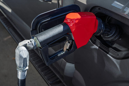 Close-up of a red fuel nozzle inserted into the gas tank of a dark gray vehicle during refueling....