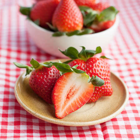 Small plate and bowl filled with succulent juicy fresh ripe red strawberries on table with red checkered tableclothの写真素材