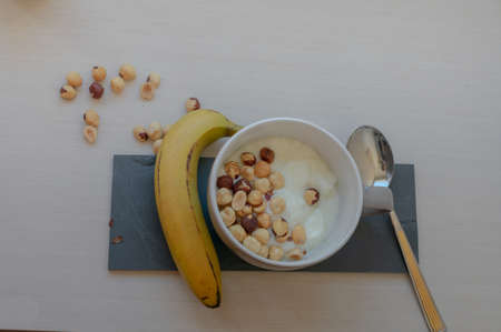 A bowl of yoghurt with hazelnuts, a banana and a spoon, with some hazelnuts on the table, top view...