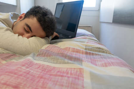 Young caucasian man tired in front of his laptop, sleeping on his bed. Overworking, saturation concepts.の写真素材