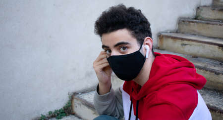 Close-up of a young caucasian man putting an earphone on his ear, while sitting on some stairs, wearing a black face mask and casual clothing.の写真素材