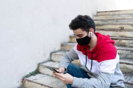 Young caucasian man wearing a black face mask and casual clothing, sitting on some stairs, with wireless earphones checking his phoneの写真素材