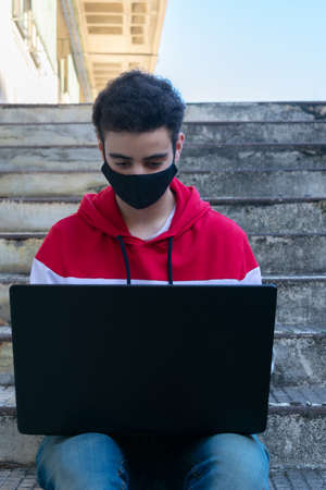 Young caucasian man, wearing casual clothes, working on his laptop, while sitting on weathered stone stairs, outdoors, with a black face maskの写真素材