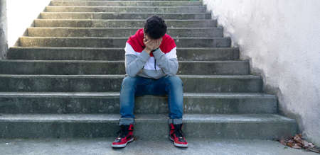 young depressed caucasian man, with his hands on his face, sitting on some stairs. Wearing casual clothing.の写真素材