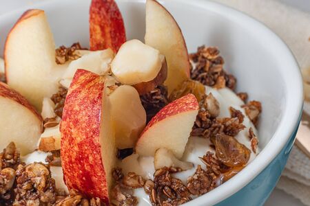 A medium blue bowl with homemade granola, plain yogurt, apple and chestnut. On white background with towel napkin and golden spoon.の写真素材
