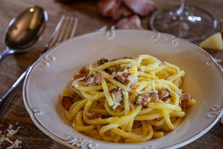 A delicious Italian Carbonara pasta, served in a deep dish, on a rustic wooden table, with grated Parmesan cheese, sliced bacon, two eggs on the side and a glass of wine.の写真素材