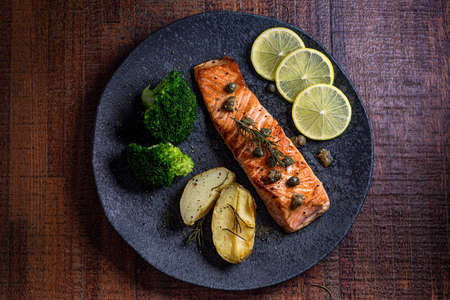 A piece of roasted salmon grilled lemon pepper and salt on a dark plate with broccoli. Wood...