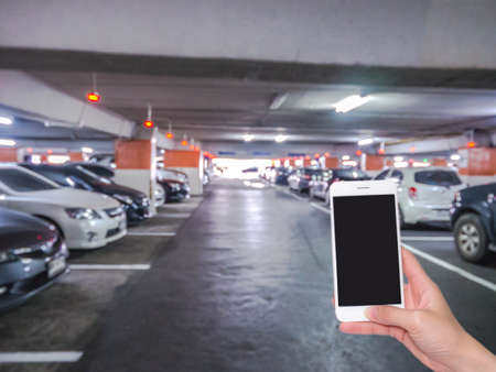 Hand holding mobile phone with indoor parking lot building blurred background and bokeh light,...