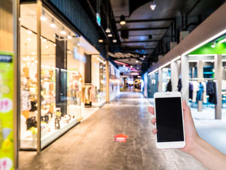 Hand holding mobile phone with Shopping mall or department store blurred background and bokeh light,...