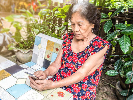 Asian senior woman unhappy while looking at mobile phone abstract loss in profits sitting in the garden, internet social media, networkの写真素材