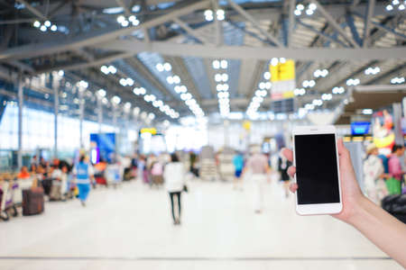 Hand holding mobile phone with airport terminal blurred crowd of Travelling people on the background, Bokeh light, Social network, internet,Traveling conceptの写真素材