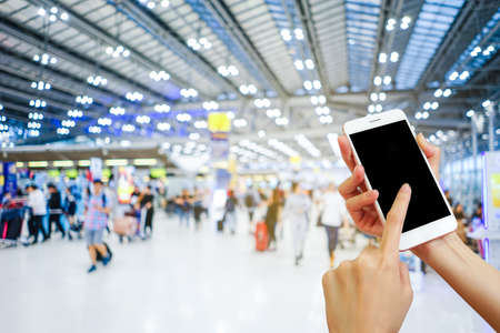 Hand holding mobile phone with airport terminal blurred crowd of Travelling people on the background, Bokeh light, Social network, internet,Traveling conceptの写真素材