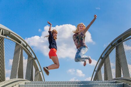 Two happy and enthusiastic teenage girls jumping on bridge in front of blue skyの写真素材