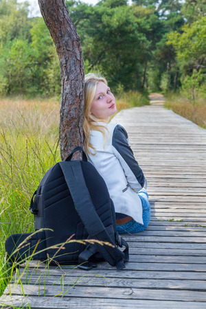 Blonde dutch teenage girl sitting on wooden path in nature with backpackの写真素材