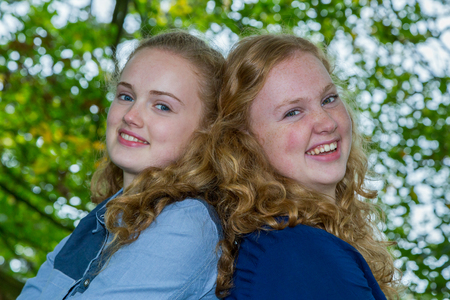 Two caucasian teenage sisters heads together laughing under green treeの写真素材
