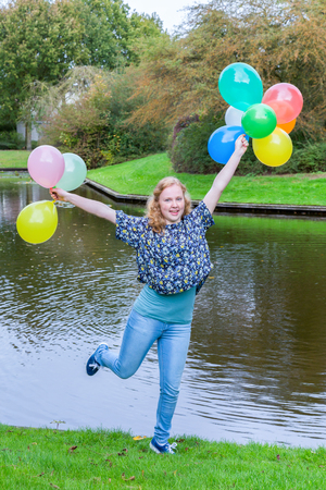 European teenage girl holding up many  coloured balloons at water of pondの写真素材
