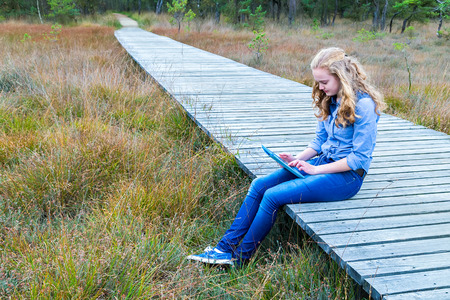 Blonde caucasian teenage girl working with tablet computer on wooden path in natureの写真素材