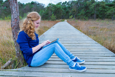 Caucasian teenage girl working with tablet computer on wooden footpath in natureの写真素材