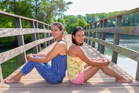 Two young women sitting together on wooden bridge over waterの写真素材