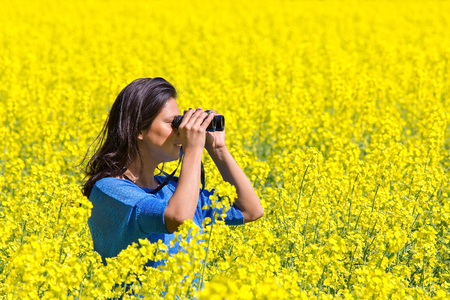 Young woman looking through binoculars in flowering yellow rapeseed fieldの写真素材