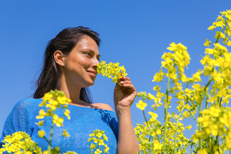 Young colombian woman smells yellow flower in rapeseed field with blue skyの写真素材
