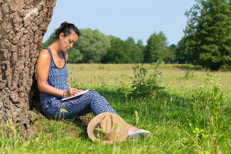 Young indian woman sitting against tree trunk writing in pastureの写真素材